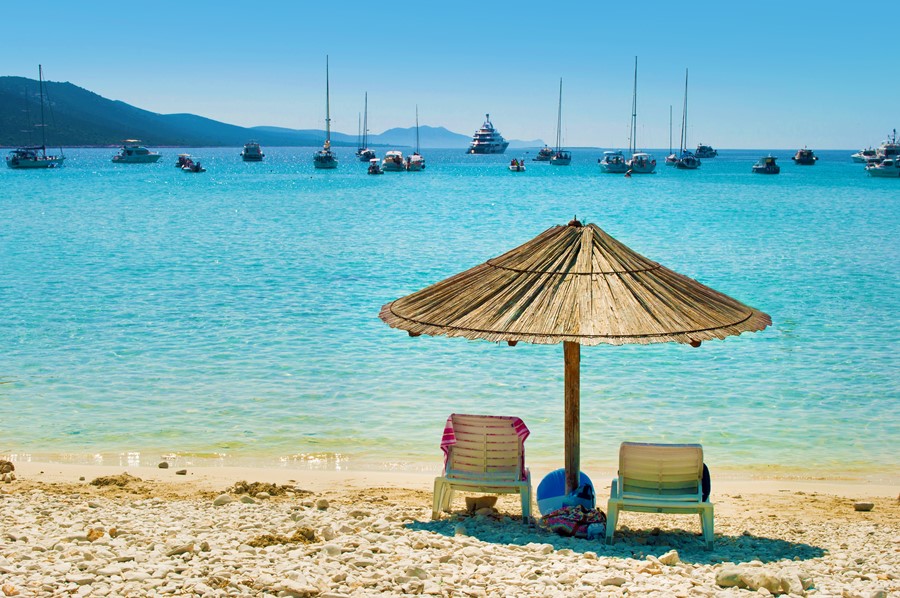 One yellow straw umbrella and two chairs on the pebble sandy beach. Many yachts in the azure blue sea lagoon in a bay near the coast of an island. Blue cloudless sky. Dugi otok Croatia